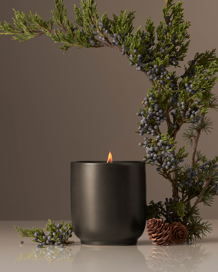 Front view of lit matte black ceramic candle with juniper sprigs and pine cones on taupe background