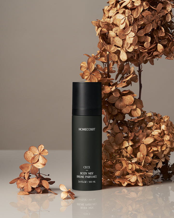 Front view of Homecourt Cece body mist, matte black cylindrical bottle with label next to dried hydrangea flowers on a beige reflective surface