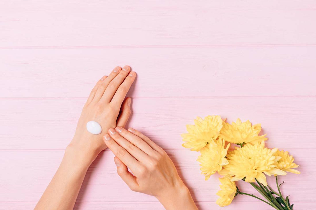 Hands applying moisturizing cream to the back of one hand beside yellow flowers on a pink wooden surface
