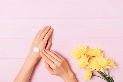 Hands applying moisturizing cream to the back of one hand beside yellow flowers on a pink wooden surface