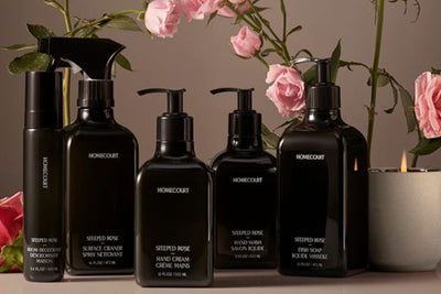 Front view of Homecourt Steeped Rose cleaning and hand care bottles showing black pump bottles with pink roses and candle background