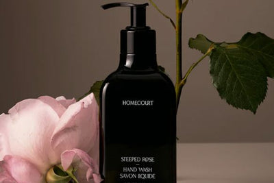 Front view of black Homecourt Steeped Rose hand wash bottle with pump beside pink rose and green leaves on neutral background
