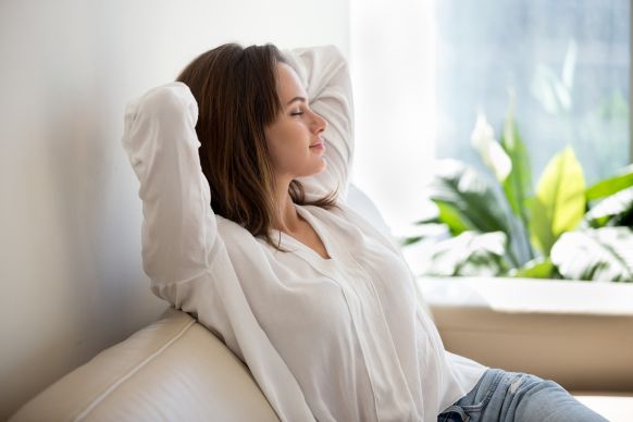 Woman leaning back on a sofa with eyes closed, relaxed in a bright living room by a sunlit window and houseplant