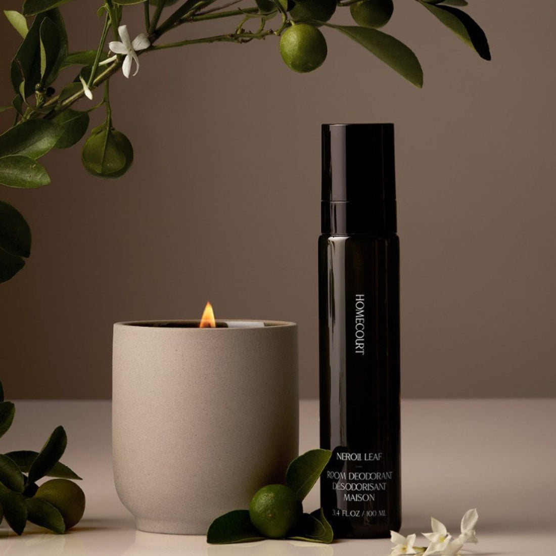 Front view of HOMECOURT Neroli Leaf room deodorant spray next to a lit ceramic candle and citrus branches on beige background