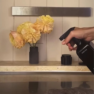 Hand spraying a black surface cleaner bottle at a countertop with a small black vase of peach-yellow flowers on the backsplash