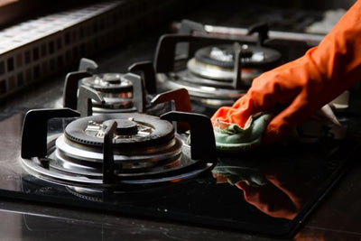 Person wearing an orange rubber glove wiping a black gas stovetop with a green cloth, cleaning around the burners