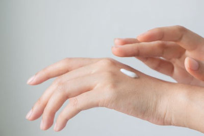 Two hands applying a dollop of white hand cream to the back of one hand against a soft gray background
