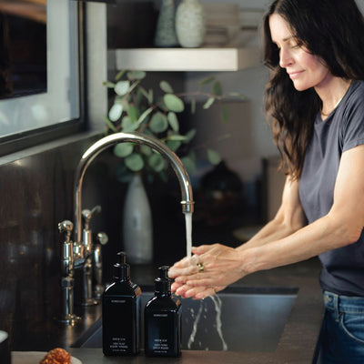 Woman washing her hands at a sink with two black hand wash bottles on the counter