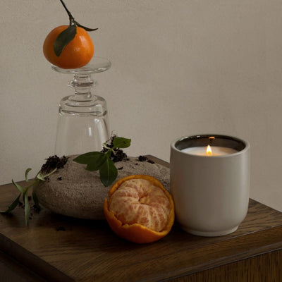 Lit candle in a white ceramic jar beside a peeled mandarin and a whole orange on a wooden table, styled still life