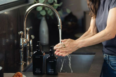 Person washing hands under a kitchen faucet with two black pump dish soap bottles on the dark countertop next to a stainless sink