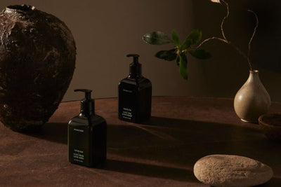 Two dark pump hand cream bottles on a brown tabletop with pottery, a smooth stone, and a green sprig in warm moody lighting