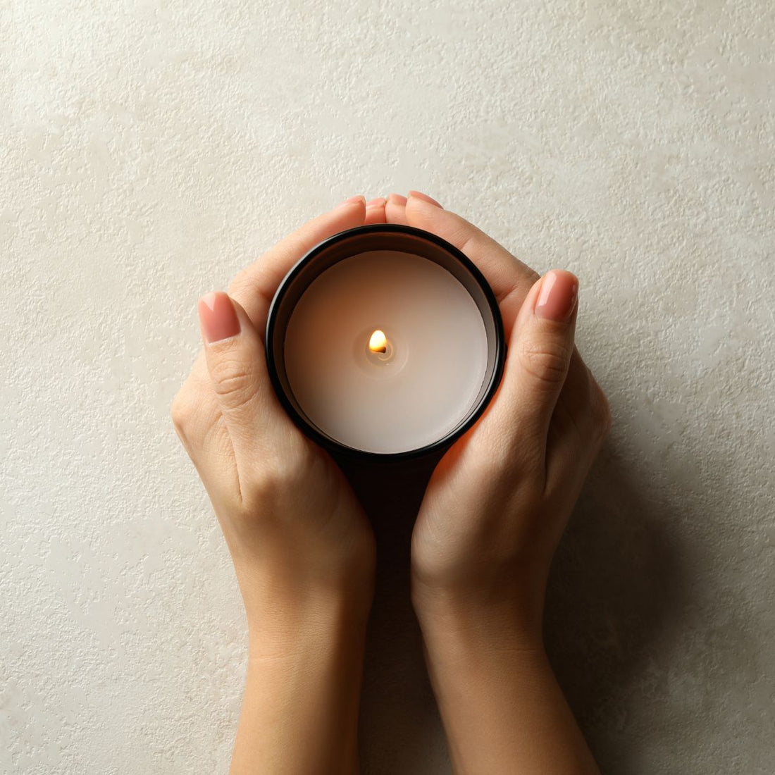 Two hands cupping a lit scented candle in a black jar against a textured beige wall