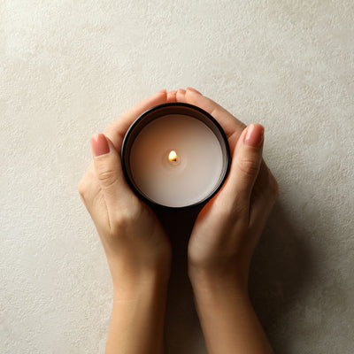 Two hands cupping a lit scented candle in a black jar against a textured beige wall