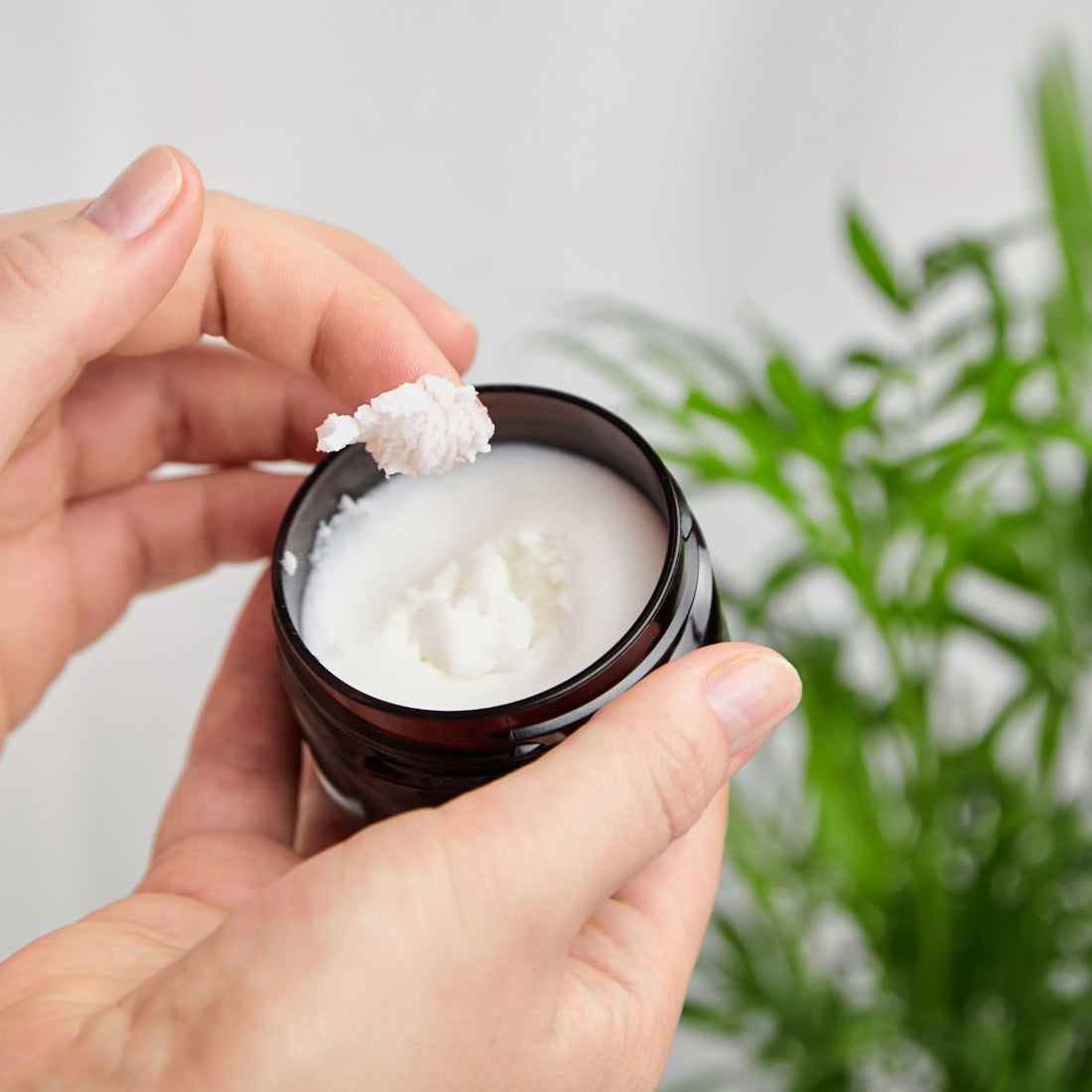 Top-down view of an open jar of hand cream showing thick white cream being scooped with hands, blurred green plant background