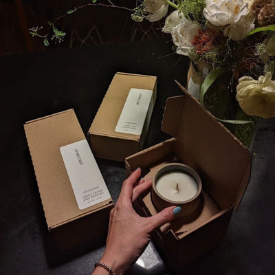 Hand holding a jar candle lifted from an open cardboard box on a dark table beside two boxed candles and a vase of flowers