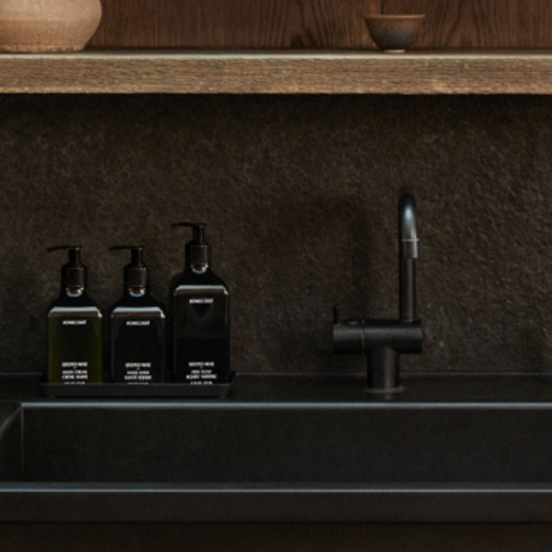 Three black pump bottles of dish soap on a tray beside a matte black kitchen sink and faucet