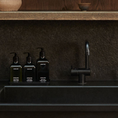 Three black pump bottles of dish soap on a tray beside a matte black kitchen sink and faucet