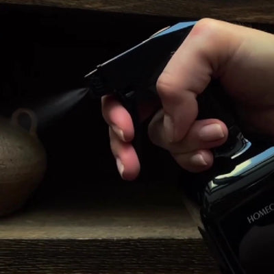 Close-up of a hand pressing the trigger on a black surface cleaner spray bottle, fine mist spraying toward a wooden shelf