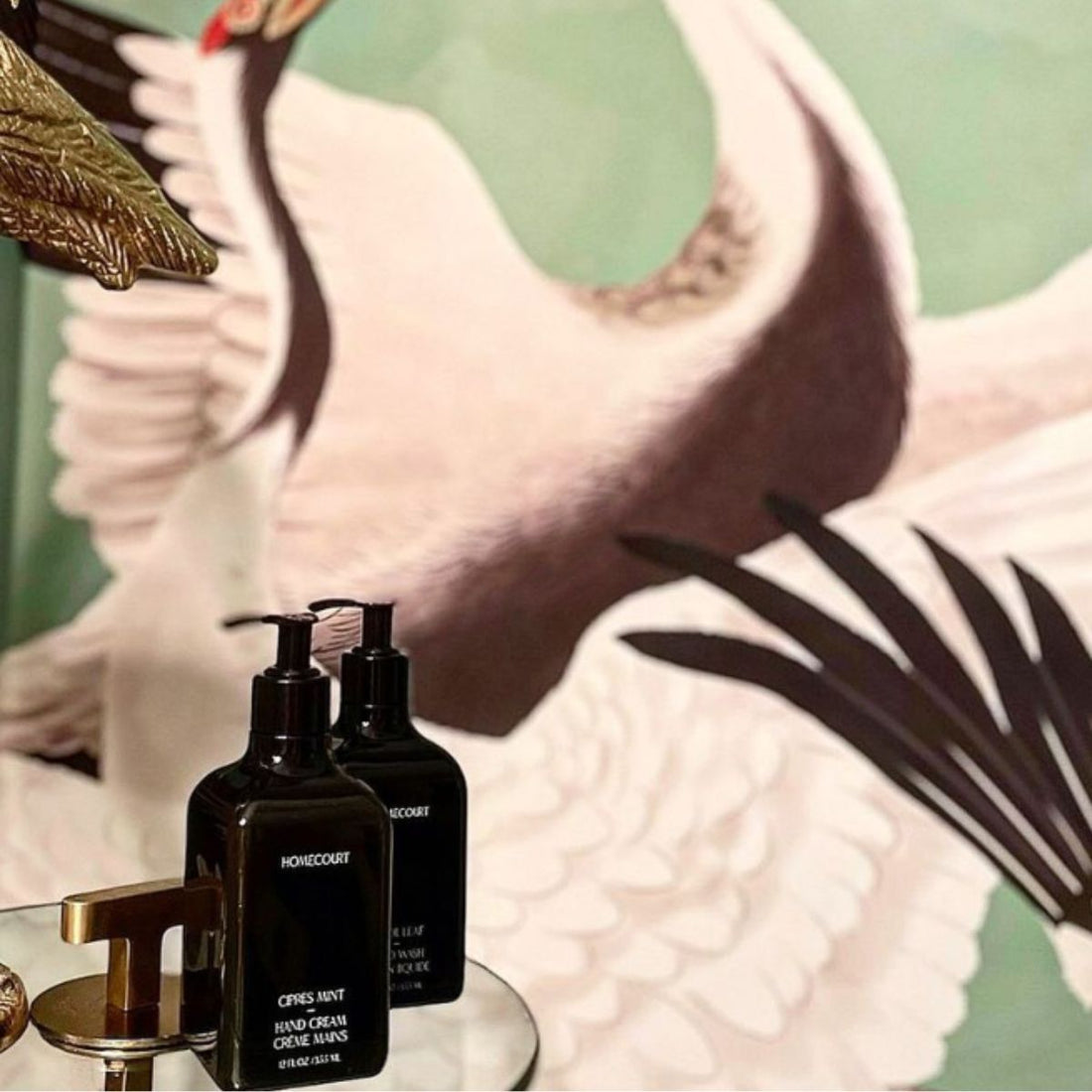 Front view of two Homecourt hand cream pump bottles on a mirrored tray with swan mural background