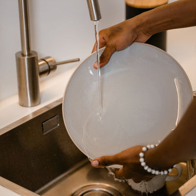 Hands rinsing a white ceramic plate under a running kitchen faucet at a stainless steel sink