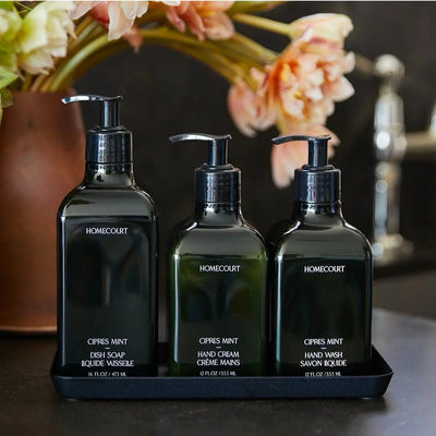 Front view of three Homecourt Cipres Mint pump bottles (dish soap, hand cream, hand wash) on black tray with blurred flowers