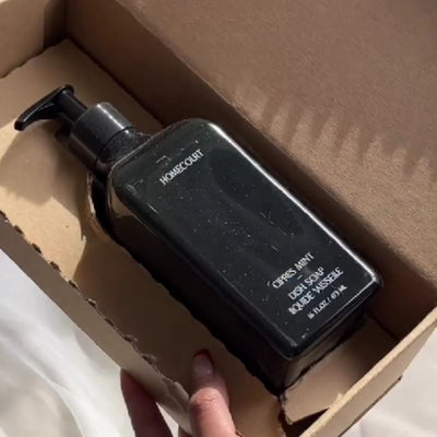 Angled top-down view of Homecourt black dish soap bottle with pump labeled ‘Citrus Mint’ inside a cardboard box held by a hand
