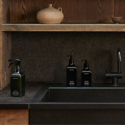 Three matte-black pump and spray bottles staged beside a matte-black sink on a dark stone countertop with a wooden shelf backdrop