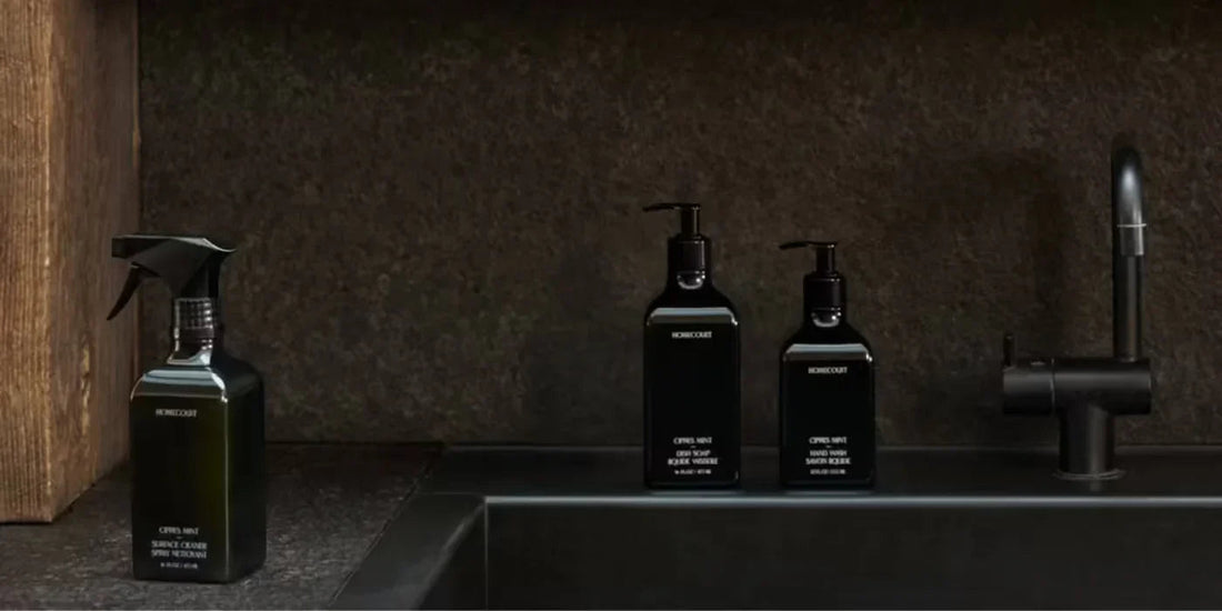 Front view of three matte black Homecourt cleaner bottles and a spray bottle with pump tops on a dark sink countertop with a black faucet