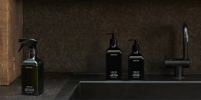 Front view of three matte black Homecourt cleaner bottles and a spray bottle with pump tops on a dark sink countertop with a black faucet