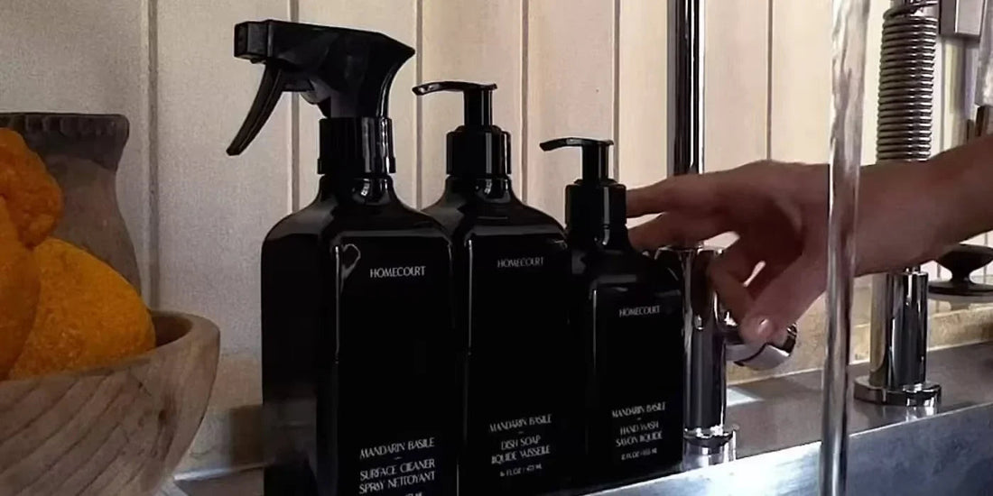 Hand reaching for a black refillable dish soap pump beside matching bottles on a stainless steel sink with a wooden bowl of oranges nearby