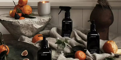 Three Homecourt cleaning bottles on draped linen with mandarins, figs, a lit candle, and rustic tabletop props