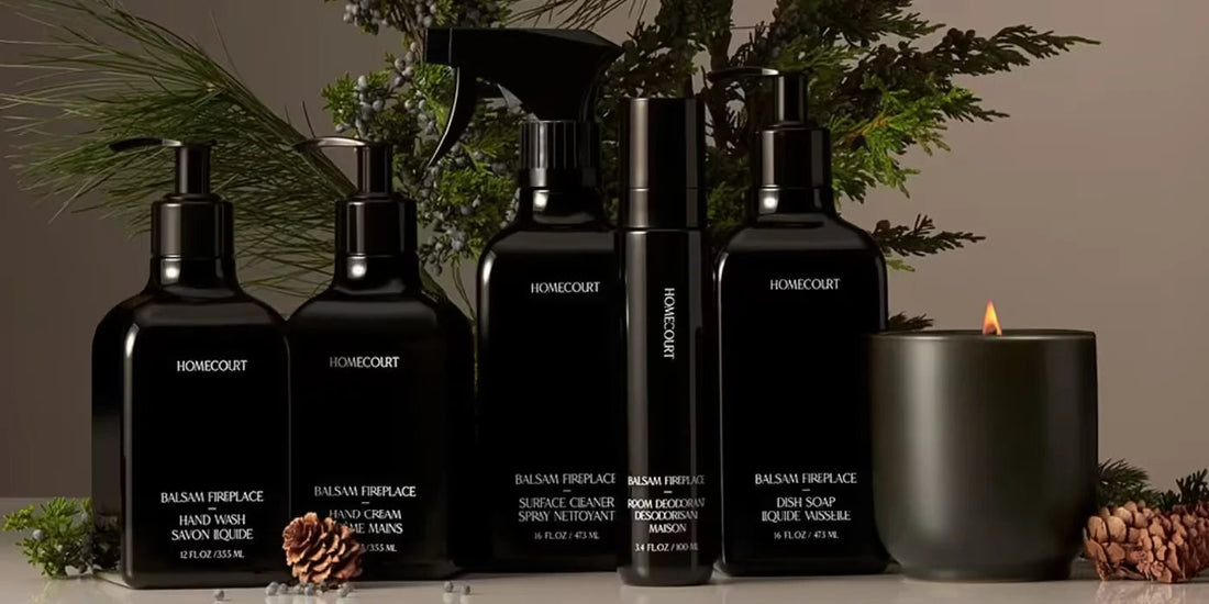 Black Homecourt dish soap and cleaning bottles arranged with a lit candle, pine sprigs, and pinecones on a neutral background