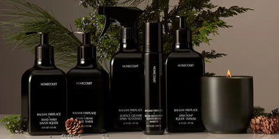 Black Homecourt dish soap and cleaning bottles arranged with a lit candle, pine sprigs, and pinecones on a neutral background