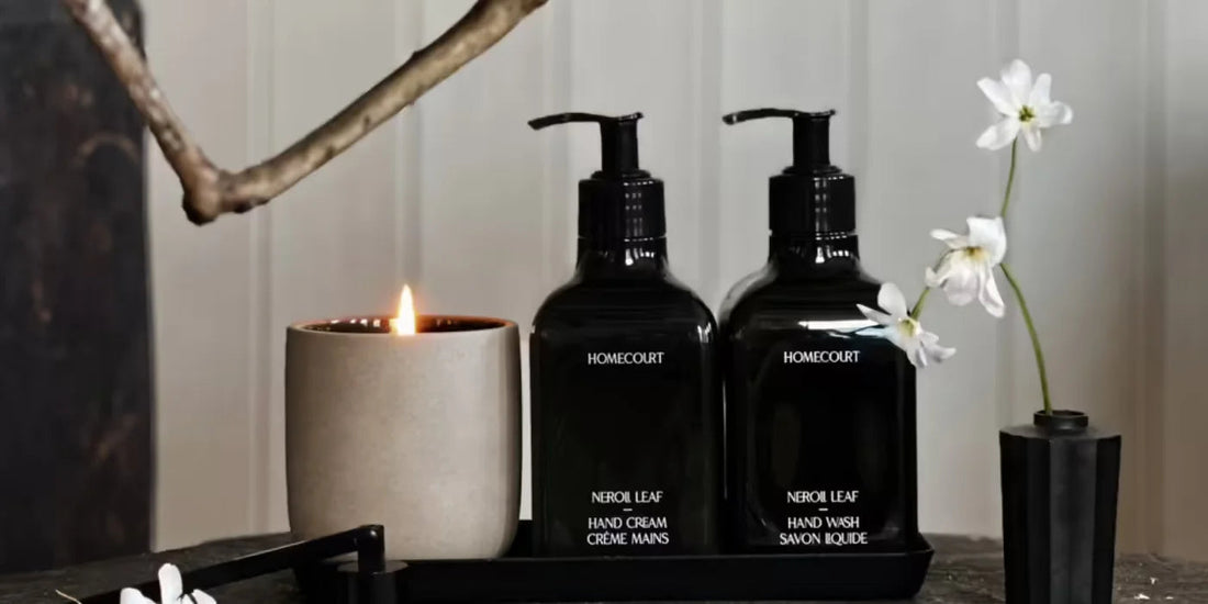 Front view of two Homecourt Neroli Leaf pump bottles (hand cream and hand wash) on a black tray with a lit candle and white flowers