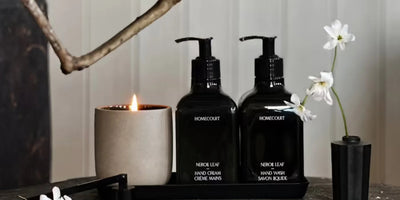 Front view of two Homecourt Neroli Leaf pump bottles (hand cream and hand wash) on a black tray with a lit candle and white flowers