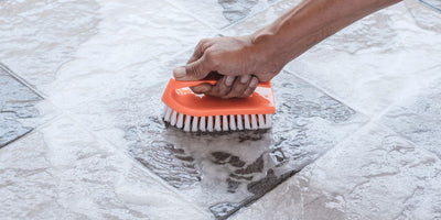 Hand scrubbing a soapy tile floor with an orange bristle scrub brush