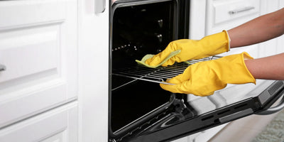 Hands in yellow rubber gloves wiping an oven rack inside an open oven in a bright white kitchen