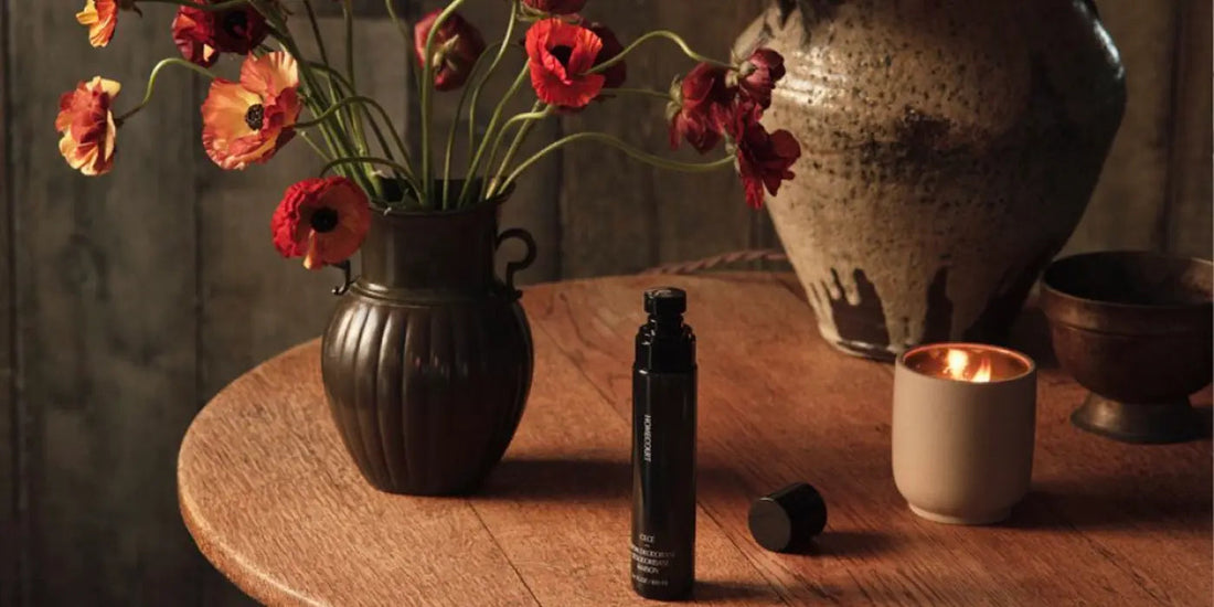 Black room deodorizer bottle on a wooden table beside a vase of red poppies and a lit candle in a warm rustic setting
