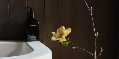 Black Homecourt hand wash bottle on a white sink beside a yellow magnolia branch against dark wood paneling