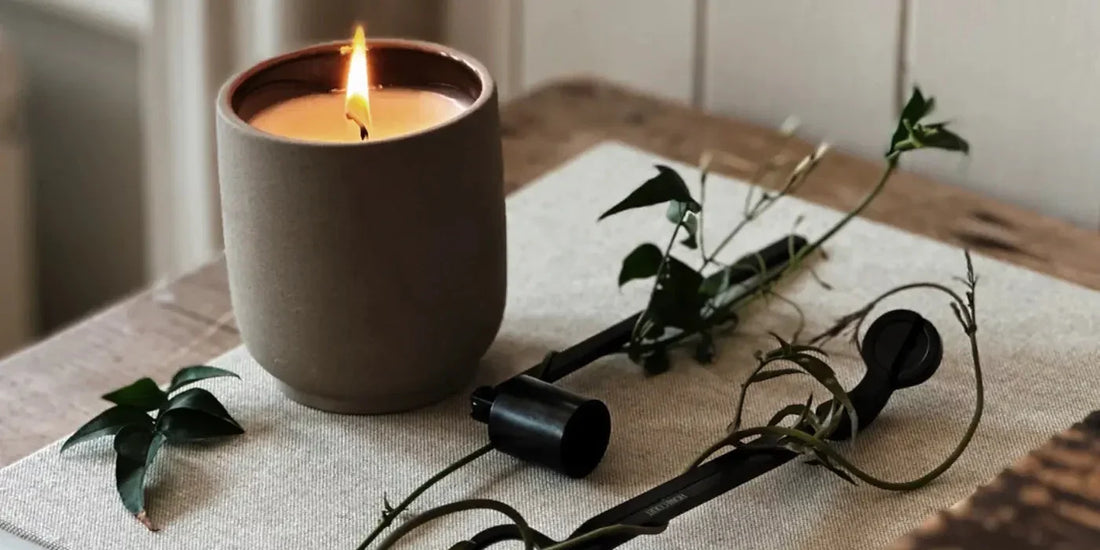 Lit candle in a beige ceramic cup on a wooden table with candle snuffer and trailing greenery, cozy neutral styling
