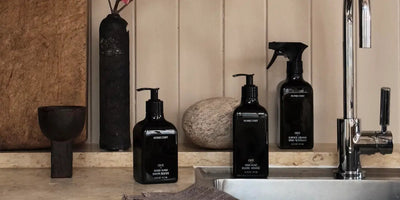 Front view of three black Homecourt pump bottles, hand wash, dish soap, and surface cleaner, on a stone sink countertop