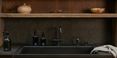 Three black hand-wash and spray bottles arranged on a dark countertop beside a matte black sink under wooden shelving with a folded towel