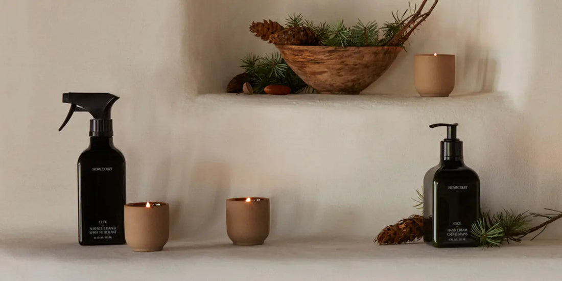 Two dark glass cleaning bottles and lit clay candles arranged on a stucco shelf with pine cones and a wooden bowl, warm natural lighting