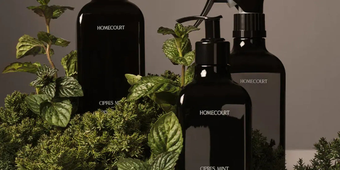 Three black Homecourt cleaning bottles surrounded by fresh mint and cypress greenery on a neutral background