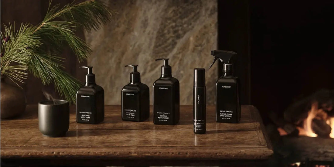 Five black Homecourt cleaning bottles lined up on a wooden table beside a lit fireplace and a pine sprig