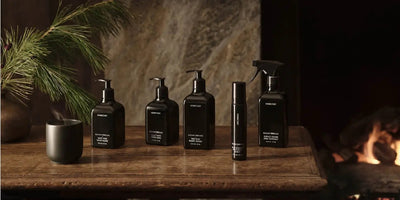 Five black Homecourt cleaning bottles lined up on a wooden table beside a lit fireplace and a pine sprig