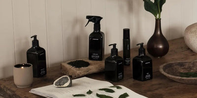 Black glass homecare bottles and a lit candle arranged on a rustic wooden table with leaves and a ceramic vase in a minimalist neutral setting