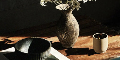 Sunlit wooden table with a rustic ceramic vase of white flowers, a textured black bowl, and a small lit candle