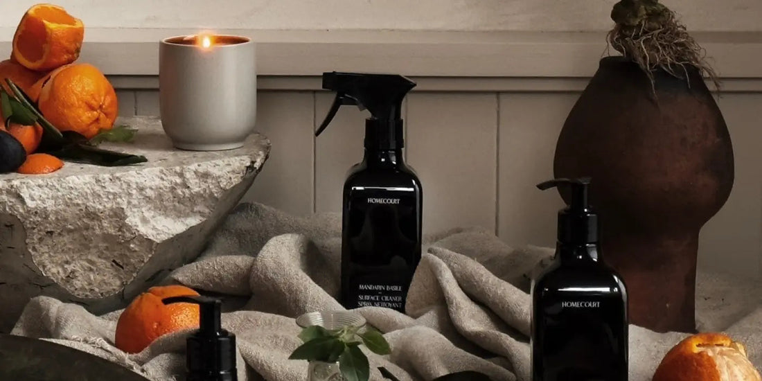 Three dark glass homecare bottles arranged on rumpled linen with a lit candle, citrus fruit, and rustic pottery in a warm tabletop vignette