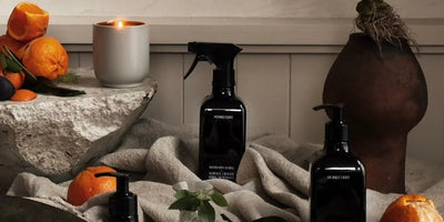Three dark glass homecare bottles arranged on rumpled linen with a lit candle, citrus fruit, and rustic pottery in a warm tabletop vignette